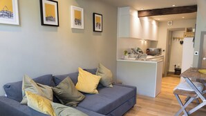 A modern living room with a grey sofa, multiple cushions, and a small kitchen area with a microwave and kettle. - The Old Middle School 1700 Cottage Addingham (Addingham)