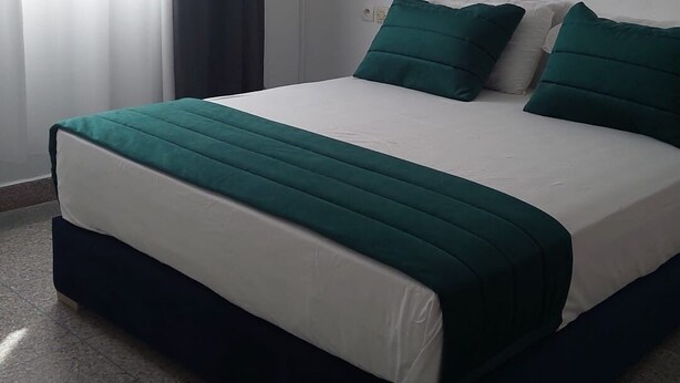 A neatly made bed with a green and white bedspread, a dark-colored headboard, and a white air conditioning unit mounted on the wall above the bed. - Large apartment with Rooftop in a private, secure, family-friendly building! (Al Aaroui)