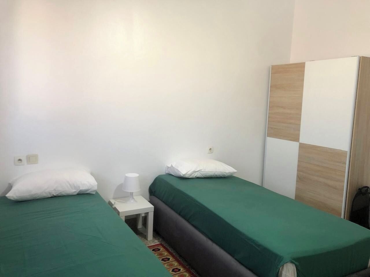 Una habitación pequeña y limpia con dos camas individuales, una mesita de noche con lámpara y un armario.