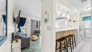 Ein modernes Wohnzimmer mit einer Couch, einem Hocker und einem kleinen Tisch mit einer Pflanze. Es gibt eine Küchenzeile mit einer Bar und Hockern.