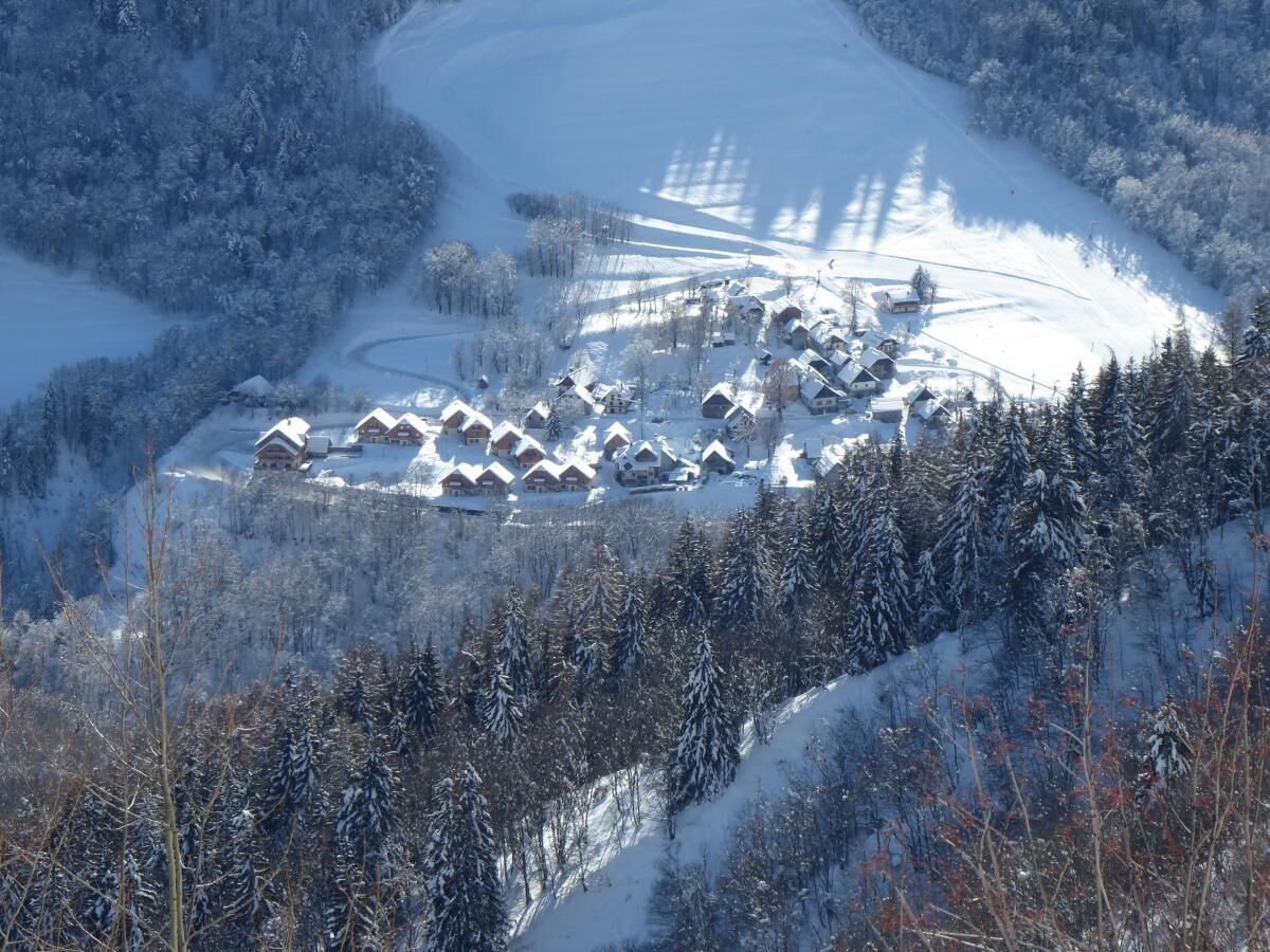 A snow-covered village with multiple chalet-style buildings clustered together, surrounded by dense forest.