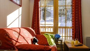Una habitación acogedora con una puerta de madera, cortinas rojas, una cama con una manta roja, una mesita con un control remoto, vasos y un libro.