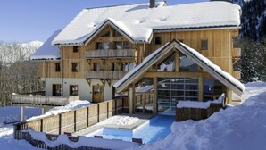 Un chalet de madera grande con varios balcones y una entrada de cristal, rodeado de nieve y montañas.