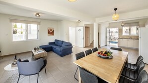 Ampla sala de estar com sofá azul, mesa de jantar de madeira com cadeiras e cozinha ao fundo.
