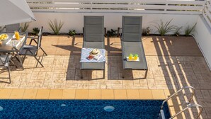 Uma área junto à piscina com espreguiçadeiras, uma mesa com bebidas e petiscos, e uma piscina com escada.