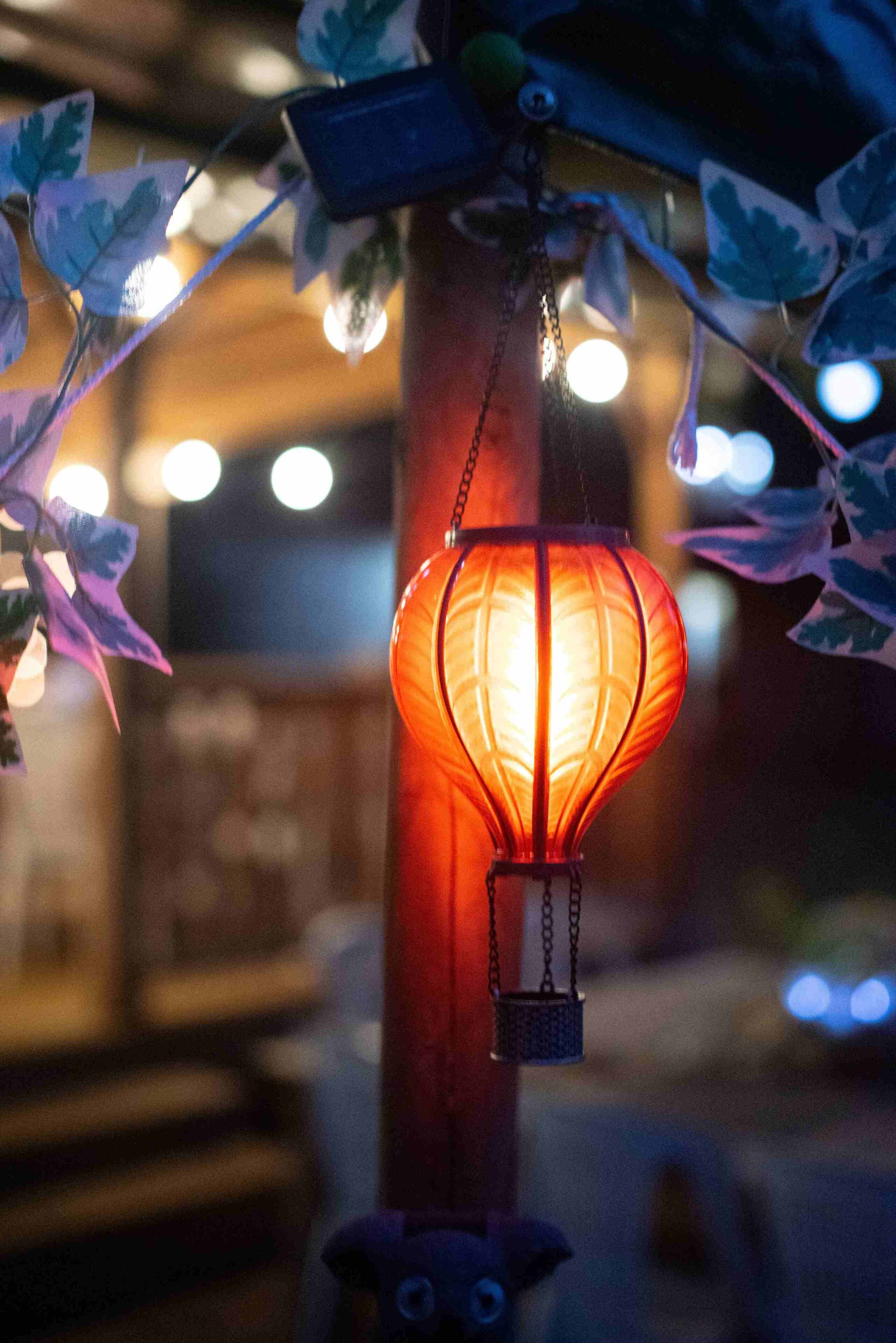 Um candeeiro suspenso com luz quente, rodeado por folhas decorativas e uma estrutura de madeira.