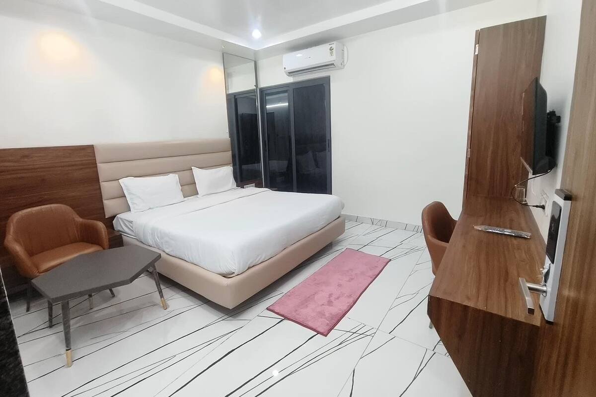 Quarto de hotel moderno com cama, duas cadeiras, uma mesinha, um espelho e televisão.