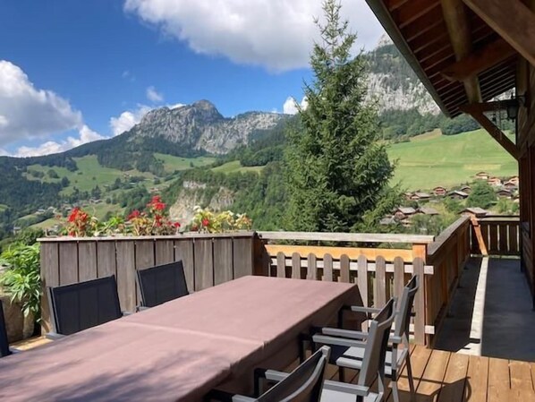 A wooden deck with a table and chairs, overlooking a mountainous landscape with houses in the valley.