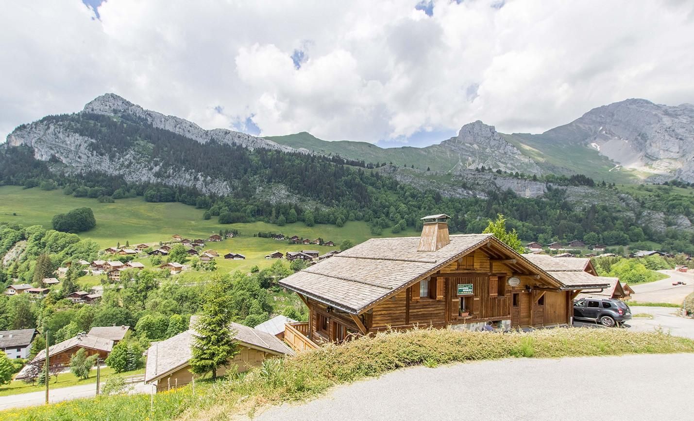 Un chalet en bois, doté d’un toit à pente, entouré d’un village rural et avec des montagnes en arrière-plan.