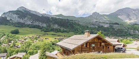 Een houten chalet met schuin dak, omgeven door een landelijk dorp en bergen op de achtergrond.