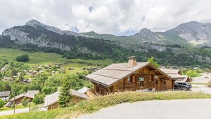 A wooden lodge with a sloped roof, surrounded by a rural village and mountains in the background. - Wifi, 50m from ski slopes, 1st floor, view mountain, terrace, balcony, fireplace or stove, parking (Le Grand-Bornand)