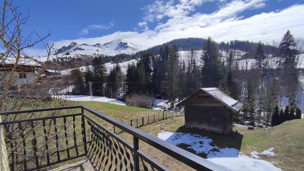 Un balcón de madera con barandilla de metal que da a un paisaje nevado, con una cabaña pequeña y montañas al fondo.