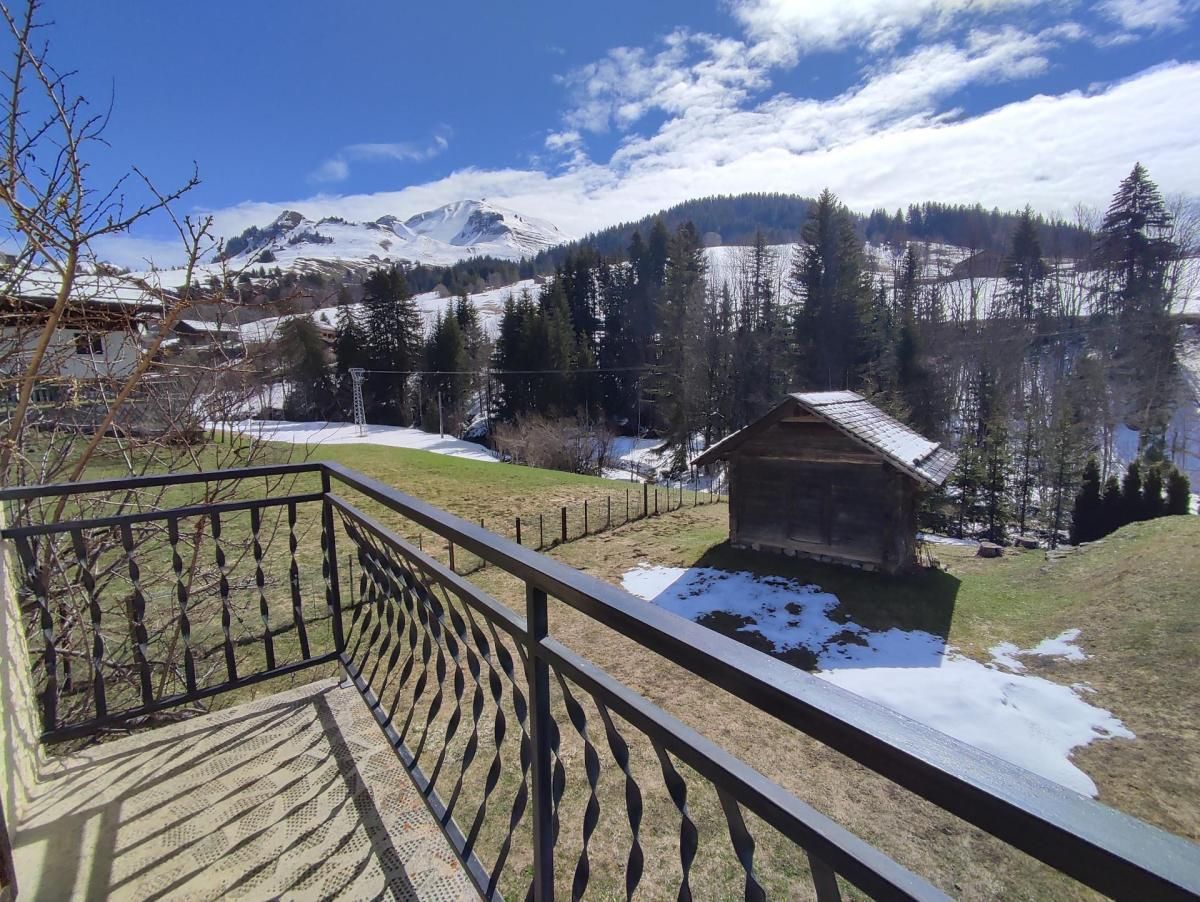 Un balcón de madera con barandilla de metal que da a un paisaje nevado, con una cabaña pequeña y montañas al fondo.