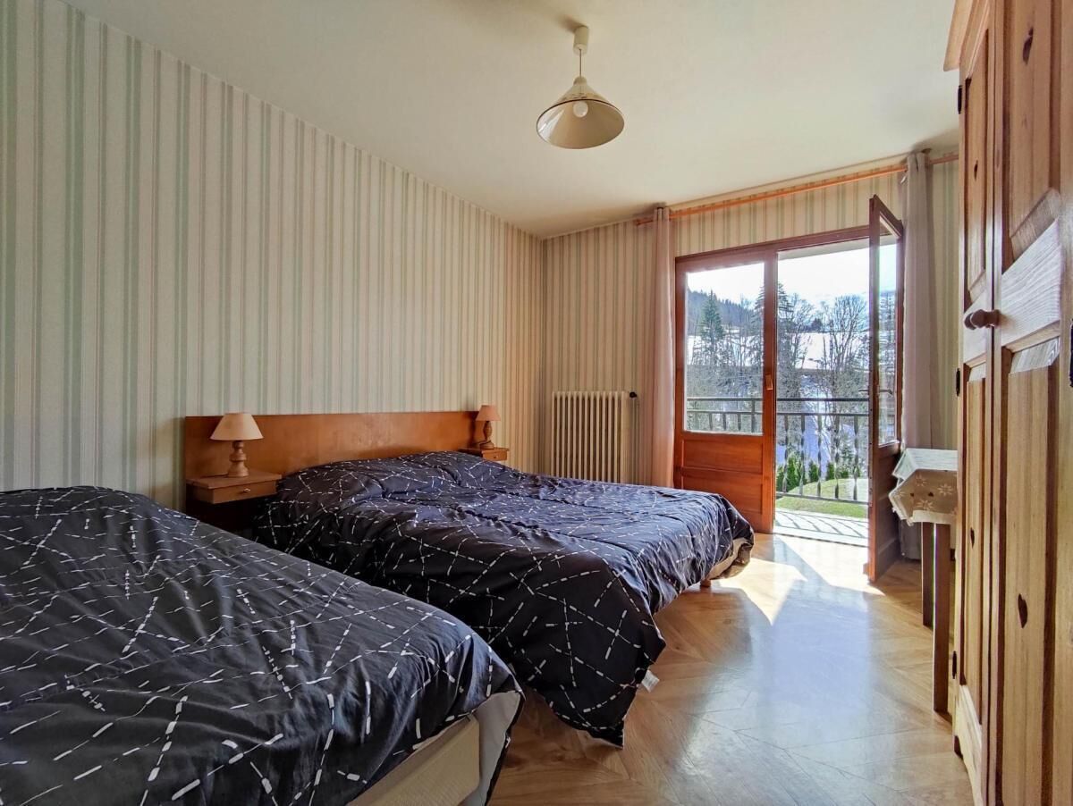 Ein Schlafzimmer mit zwei Betten, einem hölzernen Kopfstück, einem Nachttisch, einer Lampe und Blick ins Freie durch eine Glastür.