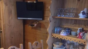 Una mesa de comedor de madera con sillas, un televisor fijado a una pared de madera y estantes con copas y platos.