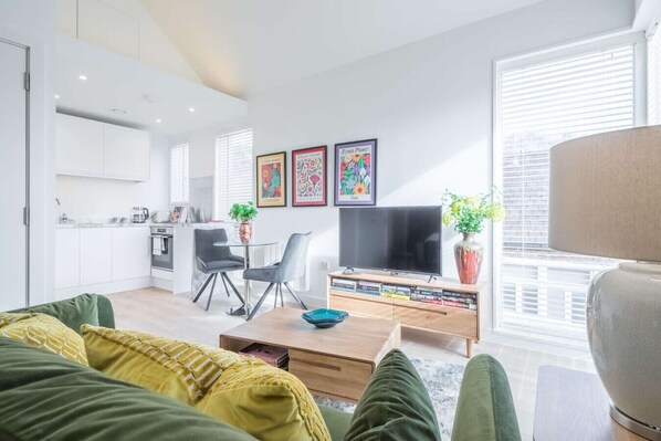 A modern living room with a sofa, a coffee table, a TV, and a bookshelf. There is a kitchen area in the background.