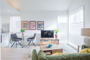 A modern living room with a flat-screen TV, two chairs, a coffee table, and a sofa. There are three framed posters on the wall and a potted plant on the coffee table.