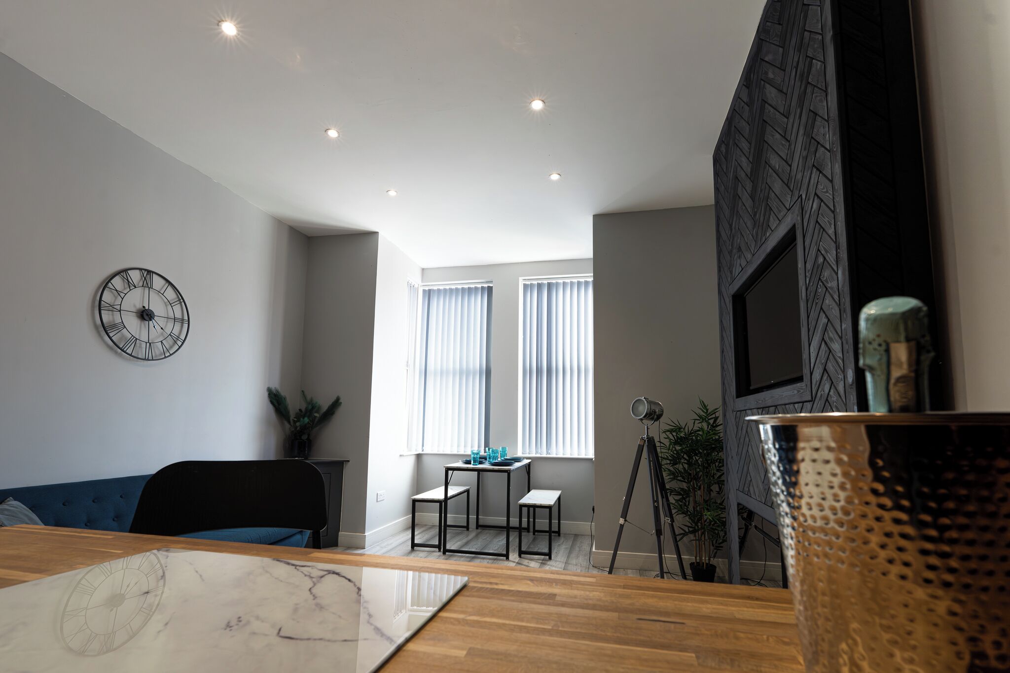 Een moderne woonkamer met een houten eettafel, een blauwe bank, een zwarte klok en een televisie aan een zwarte muur.