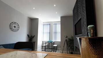 Een moderne woonkamer met een houten eettafel, een blauwe bank, een zwarte klok en een televisie aan een zwarte muur.