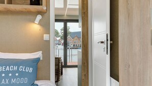 A hotel room with a bed, a nightstand, a wooden shelf, a wall-mounted light, and a door leading to a balcony with a view of buildings and water.