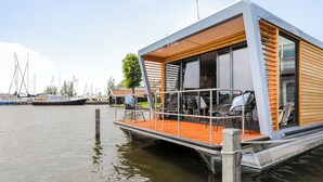 A modern floating house with a deck, outdoor seating, and a view of boats in the background.
