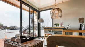 A modern room with a large window overlooking a body of water, a wicker ottoman, a desk with a chair, and a hanging light fixture.