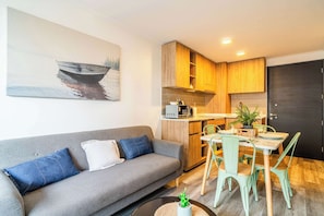 Una sala moderna con un sofá gris, una mesa de comedor con sillas, una zona de cocina con armarios de madera y un cuadro de un barco en la pared.