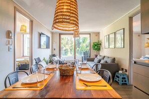 A modern dining area with a wooden table set for four, a wicker pendant light, and a view of the outdoors through large windows.