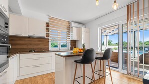 Une cuisine moderne dotée d’un îlot central, de meubles blancs, d’éléments décoratifs en bois et offrant une vue sur une piscine grâce à de grandes portes-fenêtres coulissantes.