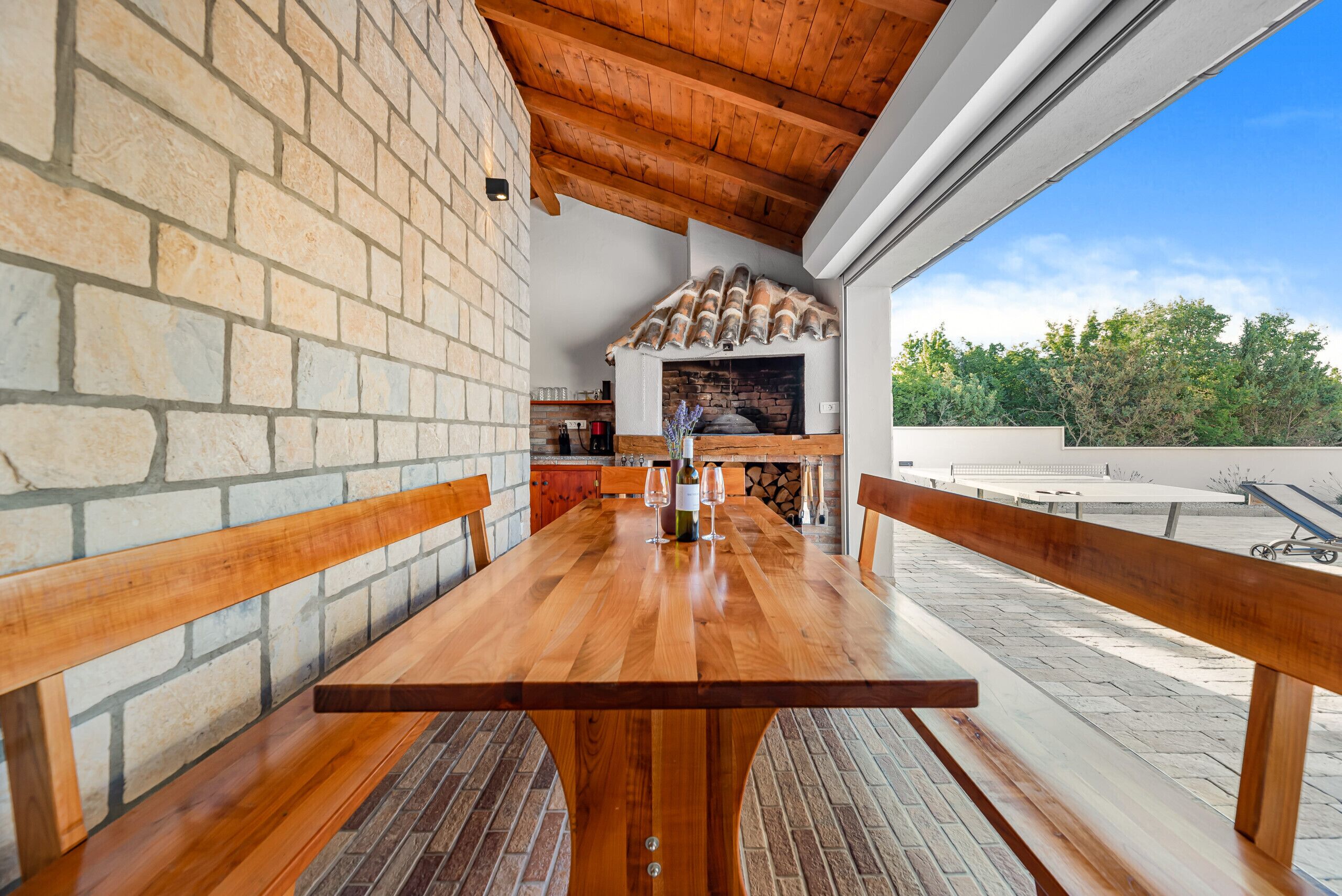 Uma mesa de madeira com bancos numa área externa coberta, paredes de pedra e forno de tijolos.