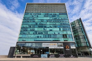 Ein modernes Hochhaus mit Glasfassade, mehreren Etagen und einer auffälligen Schanze, die eine Wohnung mit drei Schlafzimmern zum Verkauf wirbt.