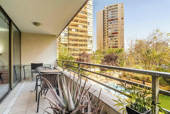 A balcony with chairs and potted plants, overlooking a swimming pool and tall buildings.