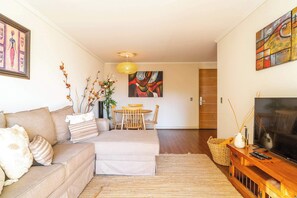Ein Wohnzimmer mit Sofa, Fernseher, einer Holzkassette, einem Esstisch mit Stühlen und Kunstwerken an den Wänden.