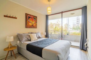 A bedroom with a bed, a painting, a lamp, a window with a view of buildings, and a balcony.
