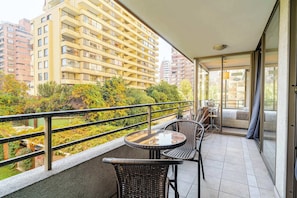 Ein Balkon mit Korbmöbeln, einem Glastisch und Blick auf ein Hochhaus und Grünflächen.