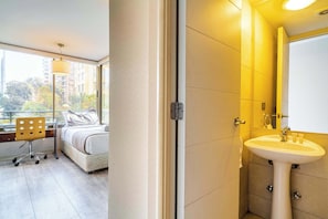 Et hotelværelse med seng, skrivebord, stol, vindue med udsigt til bygninger og badeværelse med vask og spejl.