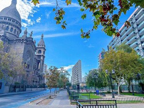 Une rue de la ville avec un grand bâtiment historique, un immeuble moderne de grande hauteur et un parc avec des bancs.