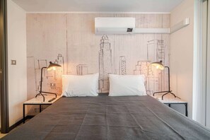 Ein Hotelzimmer mit einem Bett, zwei Nachttischen, zwei Lampen, weißen Kissen und einer Wand mit Stadtblick-Motiv.