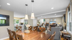 Una cocina moderna con armarios blancos, una zona de comedor con mesa y sillas de madera, y una sala de estar con sofá gris y televisor.