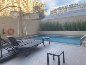 A rooftop pool area with lounge chairs and a small table, surrounded by a high wall and buildings in the background. - HOM I Depto piso 8 · Metro Santa Isabel · Gym A/A (Santiago)