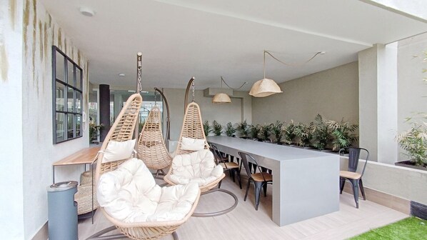 Un espacio moderno para comer al aire libre, con sillas de mimbre y una mesa larga, rodeado de plantas en macetas e iluminación colgante.