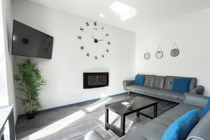 A modern living room with a large wall clock, a flat-screen TV, a fireplace, a grey sofa with blue cushions, and a coffee table.