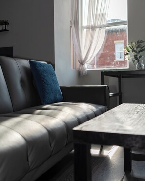A modern living room with a leather sofa, a wooden coffee table, and a view of a brick building through a window with sheer curtains.