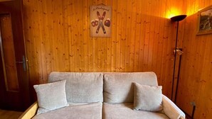 A wooden-paneled room with a beige sofa and two pillows. A wooden door is visible on the left, and a wall-mounted sign with crossed skis and snowflakes is above the sofa. - Ground floor, view village, balcony, tv, 22m², Le Grand Bornand (Le Grand-Bornand)