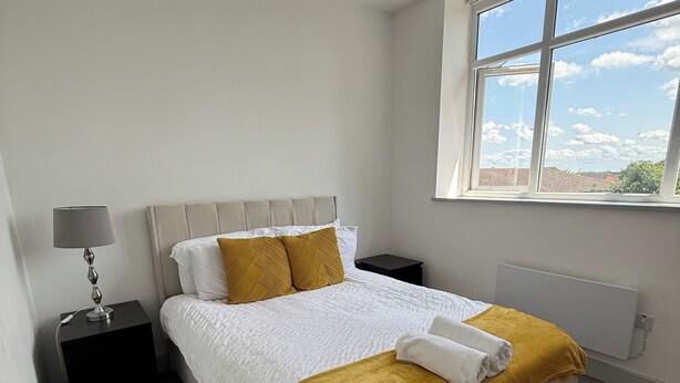 A bedroom with a bed, a nightstand, a lamp, a window, and a view of the sky. - Desborough Apartment Retreat (Kettering)