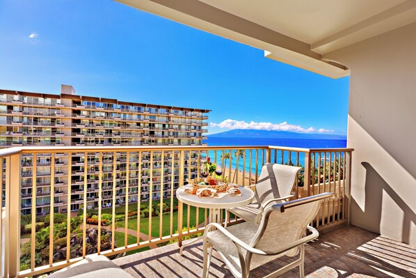 A balcony with a table and chairs, overlooking a beach and a hotel building.