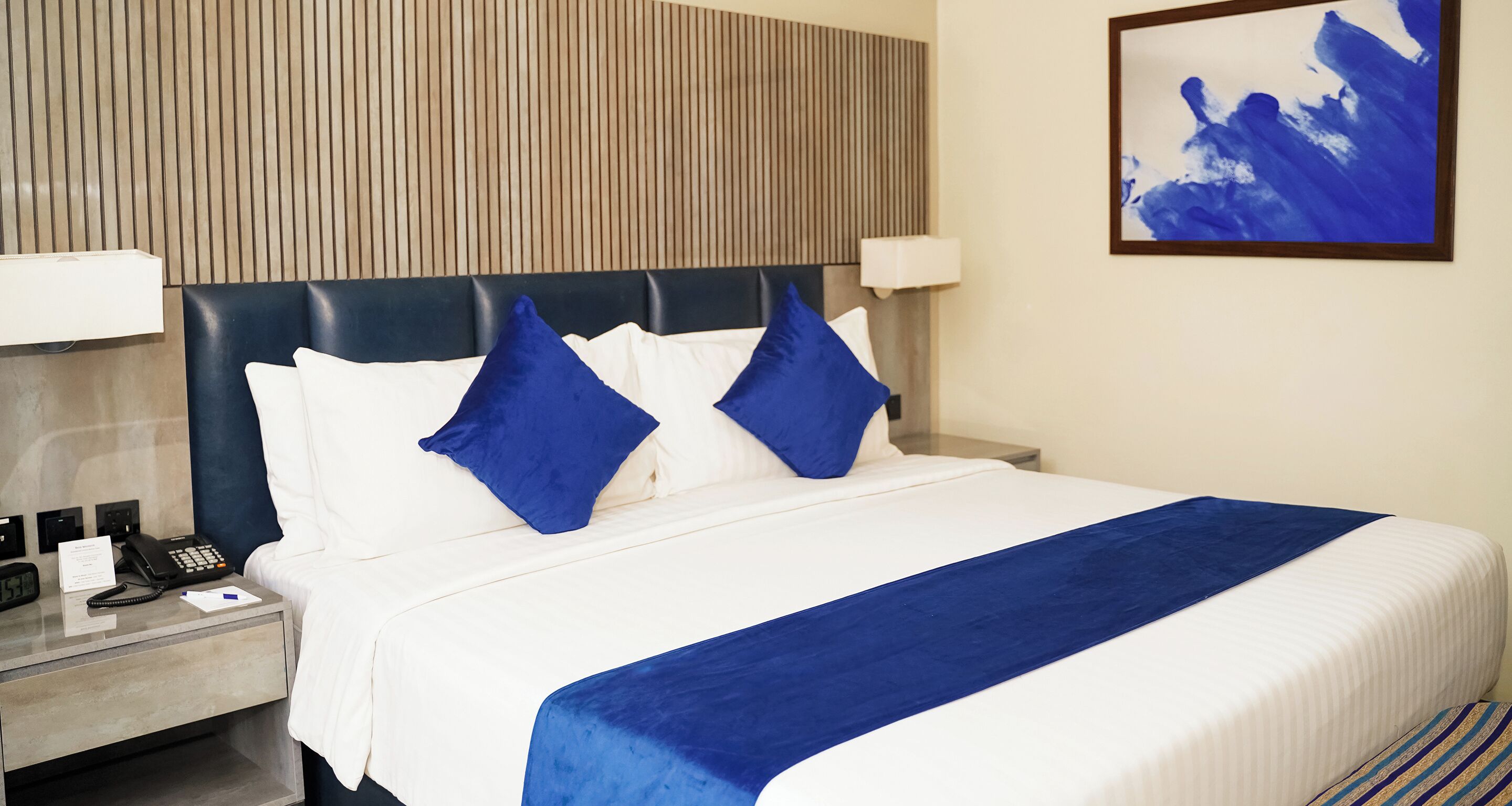 A hotel room with a bed, a bedside table with a phone, and a wall-mounted artwork.
