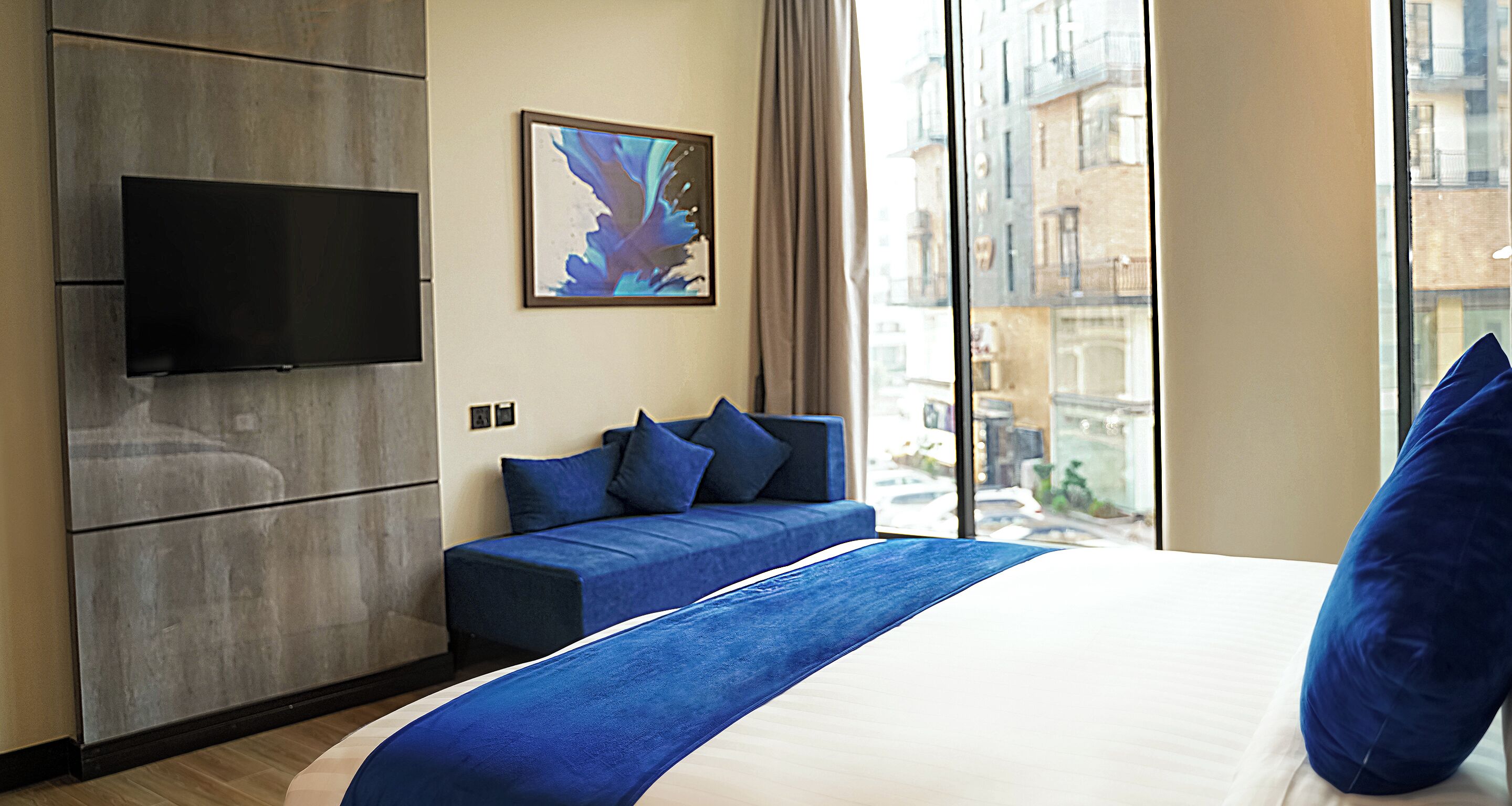 A hotel room with a bed, a sofa, a flat-screen TV mounted on the wall, and a large window offering a view of buildings.