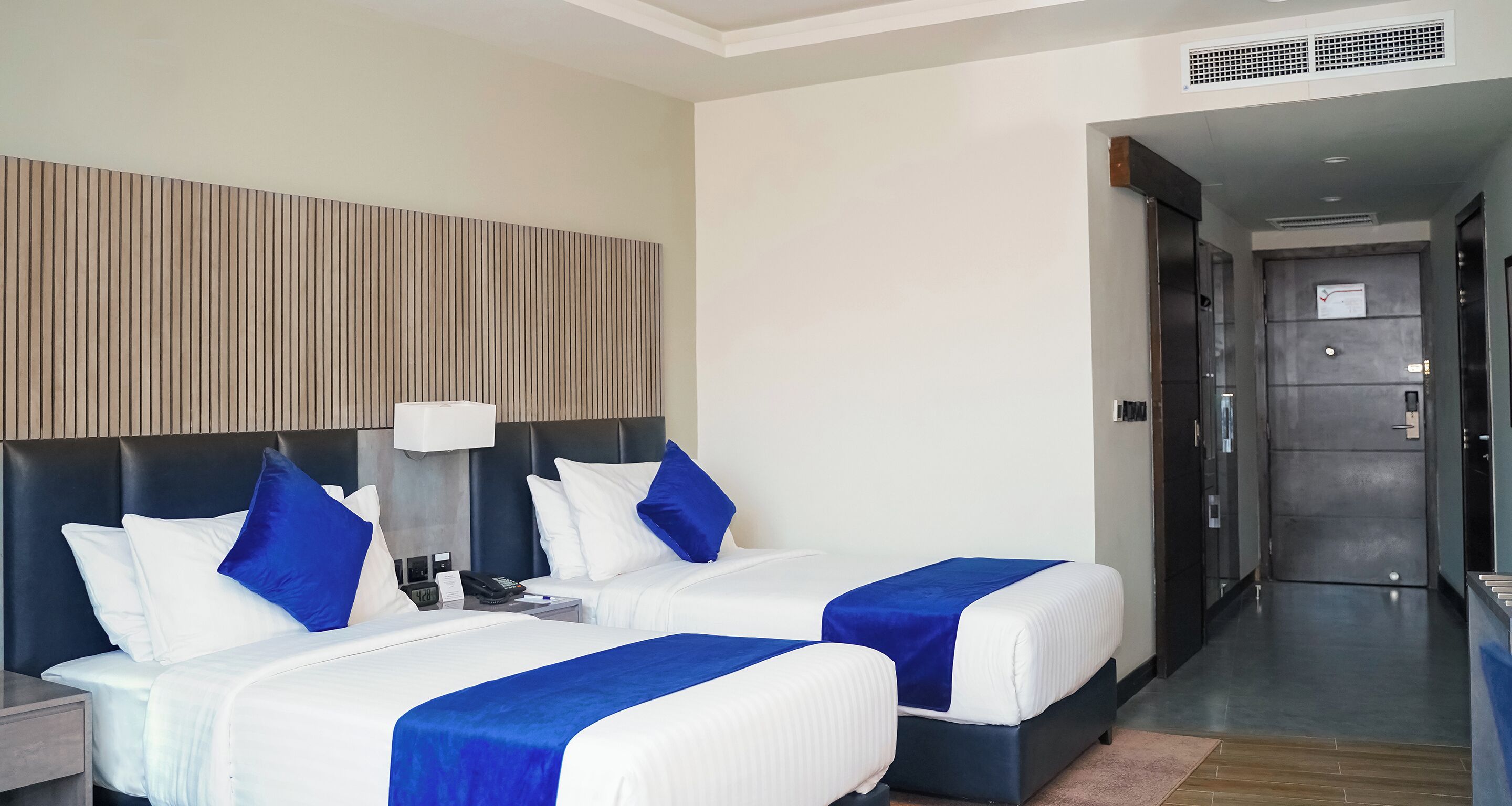 A hotel room with two beds, a bedside table, a lamp, and a wall-mounted panel.