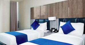 Een hotelkamer met twee bedden, elk met een blauwe sprei en witte lakens. Achter de bedden bevindt zich een wand van houten panelen, een nachtkastje met een lamp en een telefoon.